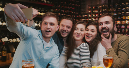 Caucasian male and female friends taking selfie photo with phone camera in bar when watching sport game. Men and women making photos on smartphone and smiling in pub. Football fans watch TV with beer.の写真素材