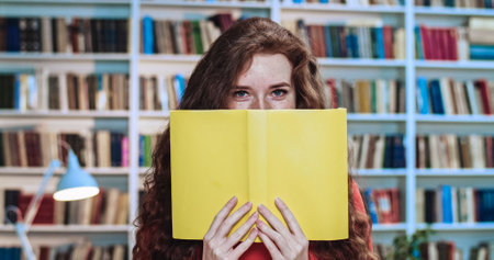 Portrait of playful red head pretty girl with long curly natural hair and yellow books having fun in...