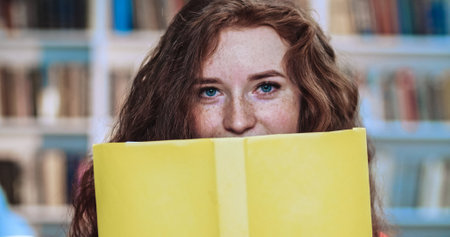 Close up portrait of sexy red head female student with long curly natural hair hiding behind yellow book in library. Bookcase in background.の写真素材