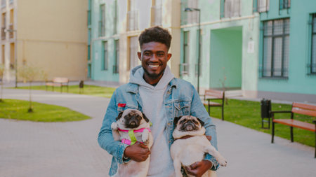 Handsome african american male in casual wear holding favourite pets while standing near modern high building. Happy young man with his cute pug doggies outdoors.の写真素材