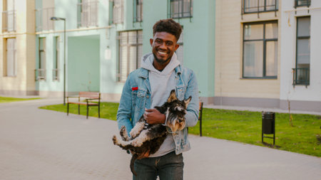 Portrait of joyful african american man dressed in casual clothes carrying his scottish terrier in hands. Happy guy smiling and looking at camera while standing outdoors with lovely pet.の写真素材