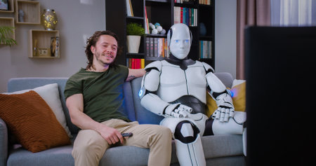 Handsome man switching channels on TV set while sitting on couch with computerized cyborg at home. Young happy male choosing video while robot making thumb up gesture.の写真素材
