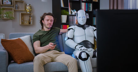 Cheerful caucasian young man with robotic cyborg watching sport game on TV while sitting on comfy sofa at home. Humanoid machine with smiling male celebrating victory on championship.の写真素材