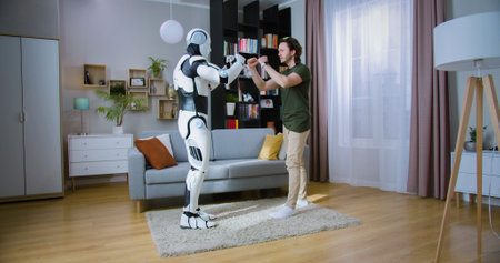 Muscular caucasian man boxing with humanoid cyborg while practicing punches in living room. Futuristic bionic robot having battle with young man at home.の写真素材
