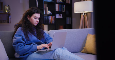 Dark haired lady working on wireless laptop while sitting on cozy couch at home in evening. Focused caucasian woman watching television while working remote in modern apartment.の写真素材