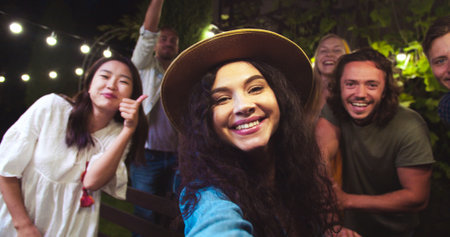 POV of happy Caucasian girl in hat talking to camera. Pretty African American and Asian girls waving hands. Handsome joyful guys smiling. Mixed-races friends at garden party in evening. Fun conceptの写真素材