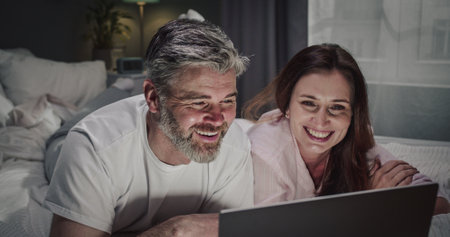 Adult woman laying at the bed with her husband and watching something funny at the laptop while spending weekend at home during the coronavirus pandemic. Couple laughing while watching movieの写真素材