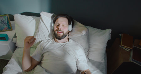 Dreamy attractive young man lying in bed, smiling and listening to music in headphones. Feeling...