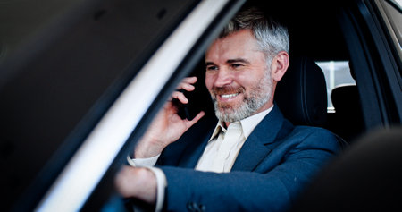 Smiling caucasian businessman talking on phone, touching steering wheel and looking around inside automobile. Successful young bearded man buy vehicle in car dealership. Close-up.の写真素材
