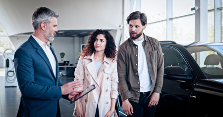 Smiling young couple discussing automobile specifications with salesman, while visiting car...