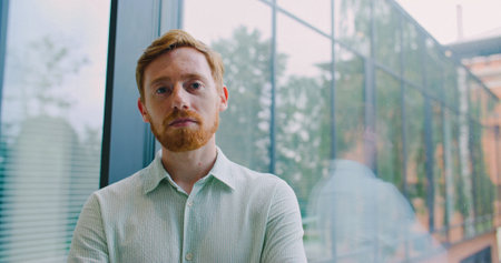 Handsome Caucasian man with red hair and beard standing in front of large glass window. Modern building. Male looking at camera with confidence and serious expression. Office building. Entrepreneur.の写真素材
