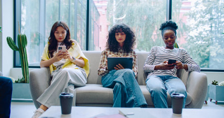Camera view of three girls sitting on couch on big resting area. Using gadgets. Scrolling...