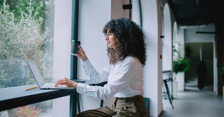 Relaxed Caucasian female sitting in front of laptop at small window ledge. Using laptop. Typing on keyboard. Entering information while relaxing during coffee break. Holding drink with one hand.の写真素材