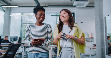 Young mixed-raced coworkers resting together during their coffee break in office. Pretty women walking in middle of room with desks and computers. Discussing about plans for weekend or working tasks.の写真素材