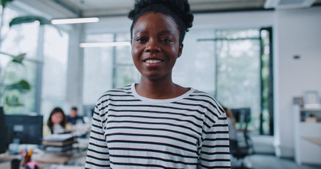 Zooming in on adorable African American woman with casual clothes standing before desks with computers. Young girl glancing with joy to camera. Expressing good feelings. In background people working.の写真素材