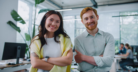 Two people standing side by side and confidently looking directly at camera. Woman with dark hair and yellow sweater over her shoulders positively smiling. Handsome man with red hair crossing his armsの写真素材