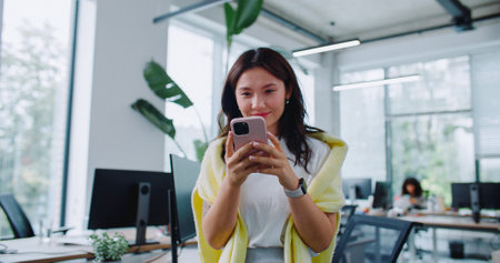 Camera moving around happy Asian woman with casual clothes spending time with phone. Young girl webbing social media or texting with someone on Internet. Using digital gadget in office. Messaging.の写真素材