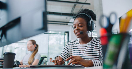 Camera view of diverse women sitting in front of computer displays. Worker on right side and wearing headset for remote consultation with clients. Working in call center. Customer support.の写真素材