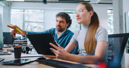 Intelligent Caucasian employees sitting together at table and discussing about new project. Handsome man with coworker comparing data from clipboards. Entering important information on computer.の写真素材