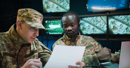 Young African American man dispatcher in headset working on laptop computer in control room. Multiethnic soldiers sitting at table and studying secret documents of military operation and strategy.の写真素材