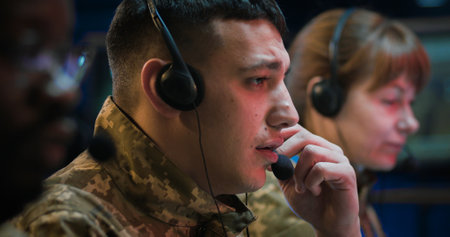 Close up of Caucasian male officer in headset working as dispatcher in monitoring room. Soldier answering calls on hot line of army controlling center. Multiethnic male and female co-workers beside.の写真素材