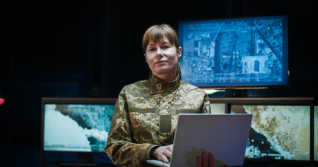 Portrait shot of Caucasian beautiful young woman from army standing in monitoring room and using laptop computer. Female soldier looking straight and smiling to camera.の写真素材