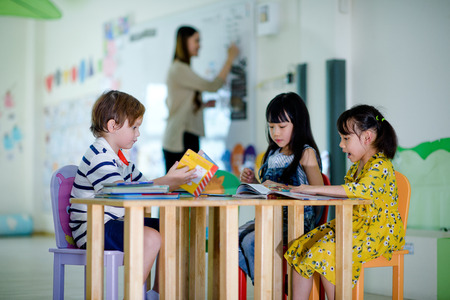 group of international kids in preschool enjoy reading books with teacher watching in backgroundの写真素材