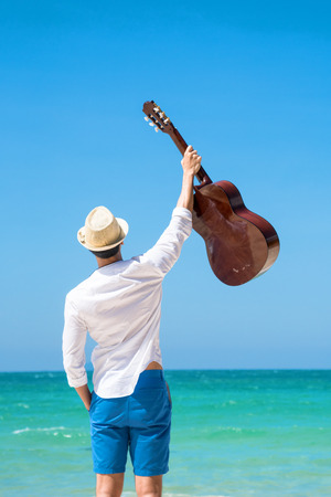 happy man holding guitar hand up in the air at sea beach with blue cleared sky in backgroundの写真素材