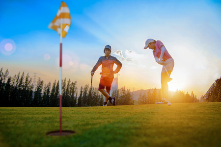 golf player looking competitor opponent putting golf ball to the hole on the green in golf course, at sunset light in backgroundの写真素材