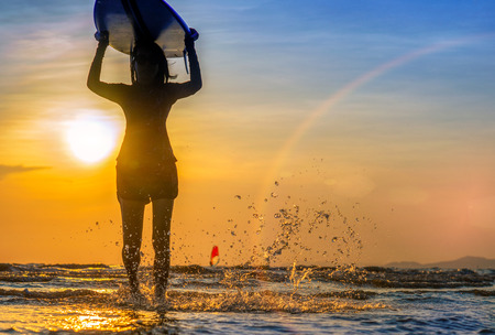 Silhouette of woman holding surfboard turn on the head walking on the edge of sea wave, splash of water kicking while walking throughの写真素材