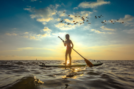 silhouette of a little girl standing on pad of surfboard holding paddle try to row scull surfboard...