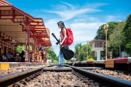 woman traveller holding guitar with backpack walks across railway at railway station to find incoming train for destrination in holiday vacation touringの写真素材