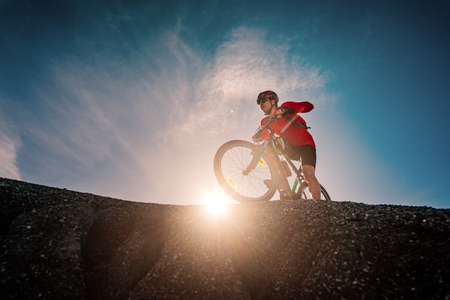 man cyclist biker riding mountain bike running from the hill at light sunset in backgroundの写真素材