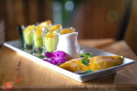 Ripe mango with sticky rice in small cups glass, topping with fresh coconut milk, prepare in white plate on green banana leaf, delicious thai dessertの写真素材