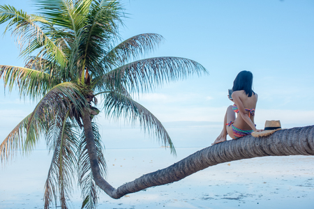 woman in bikini enjoy the seabeach by sitting onto the laying of coconut plam tree over the sea water in daylightの写真素材