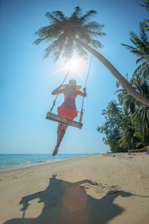 young woman in bikini tourist enjoy the sea beach nature by sitting on the wooden swing at sea beach under coconut palm tree, summer and vacation or long weekend cheerfully on the islandの写真素材