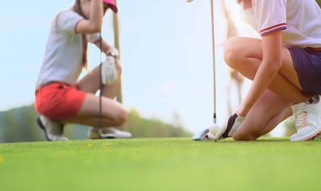hand of young woman golf player holding golf ball laying on wooden tee, prepare and ready to hit the ball to the destination target, opponent competitor or golf mate buddy watching in backgroundの写真素材
