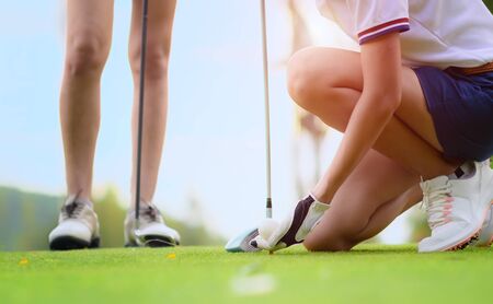 hand of young woman golf player holding golf ball laying on wooden tee, prepare and ready to hit the ball to the destination target, opponent competitor or golf mate buddy watching in backgroundの写真素材