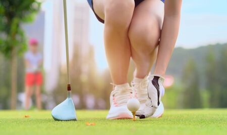 hand of young woman golf player holding golf ball laying on wooden tee, prepare and ready to hit the ball to the destination target, opponent competitor or golf mate buddy watching in backgroundの写真素材