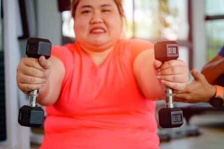 hands of fat plump woman holding dumbbell try to life up under is struction of personal trainer or coach, workout in gym for weight body reductionの写真素材