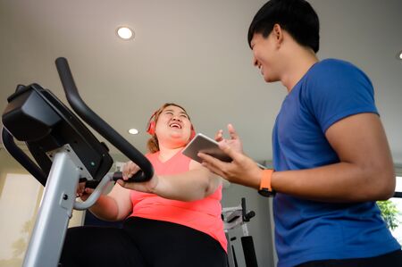 fat woman being ride on machine, workout under instruction of personal coach besideの写真素材