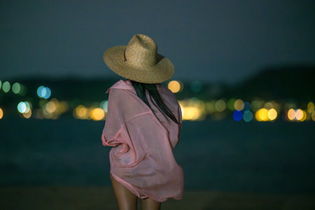 Woman stands alone at the edge of the sea beach with city lights in the backgroundの写真素材