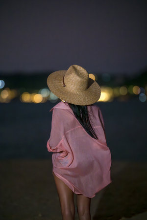 Woman stands alone at the edge of the sea beach with light of the city in backgroundの写真素材