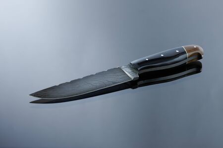 an all-metal, middle kitchen knife lies on a mirror surface, a blade made of multilayer damask...