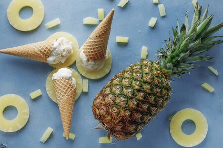 pineapple ice cream in the horn are on the pineapple rings, in the frame are three horns, not peeled pineapple, pieces of pineapple, textured blue table, top view.の写真素材