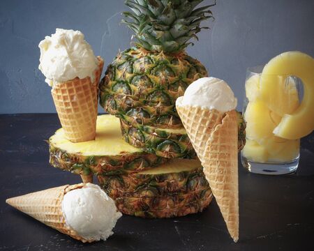 pineapple ice cream in the horn stands on a dark table against the background of a light wall, in the frame is pineapple, in the background is a glass with pineapple slicesの写真素材