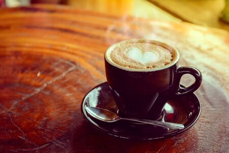 Hot coffee, heart shape with milk latte on top of black ceramic cup with metal spoon on wooden table in coffee shop and cafe in vintage tone, copy space for your text. Times to relax with caffiene concept.の写真素材