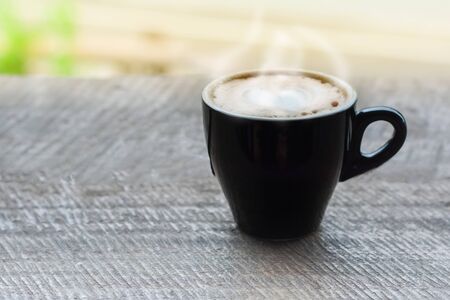 Hot coffee with smoke, heart shape with milk latte on top of black ceramic cup on wooden table in coffee shop and cafe in vintage tone, copy space for your text. Times to relax with caffiene concept.の写真素材