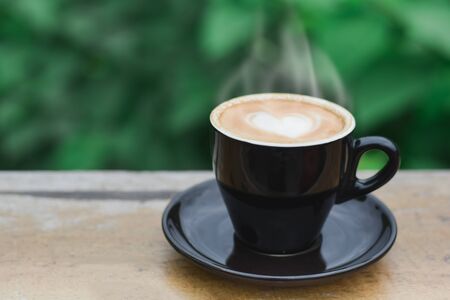 Hot coffee with smoke, heart shape with milk latte on top of black ceramic cup on wooden table in coffee shop and cafe with green background, copy space for your text. Times to relax with caffiene concept.の写真素材