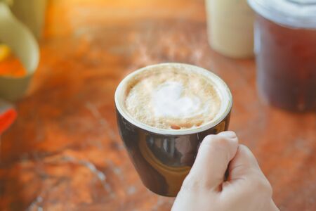Hold hot coffee, heart shape with milk latte on top of black ceramic cup on hand at coffee shop and...